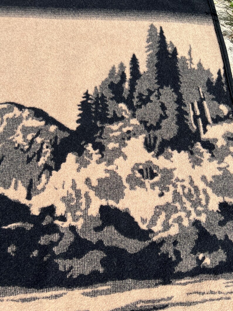 Puede incluir: Una manta tejida con un patr&oacute;n de paisaje en alto contraste negro y beige. El dise&ntilde;o muestra un bosque denso de pinos frente a picos monta&ntilde;osos escarpados, con un borde negro s&oacute;lido en la parte superior y un acabado de tela texturizada.