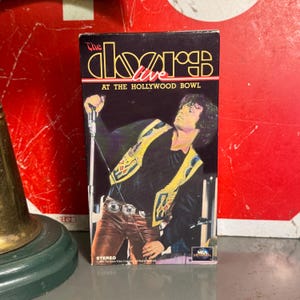 May include: A VHS tape of The Doors Live at the Hollywood Bowl. The cover features a live performance image of the band's lead singer, wearing a black shirt and a yellow vest with gold accents. The tape is in a black case with red and yellow text.