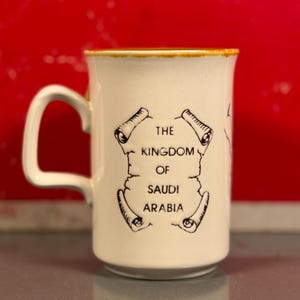 May include: Cream-colored ceramic mug with a large handle and a slightly distressed rim. The mug features the text "THE KINGDOM OF SAUDI ARABIA" in black, framed by a scroll design. The background is a solid red.