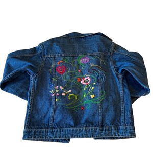 May include: A blue denim jacket with colorful floral embroidery on the back. The embroidery features red, pink, purple, and yellow flowers with green vines and leaves. The jacket has a classic collar and button-up front.