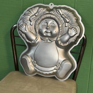 May include: A silver-colored metal cake pan in the shape of a doll with a smiling face, bows, and pigtails. The pan has a scalloped edge and a loop for hanging. The doll is in a seated position with arms outstretched.