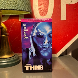 May include: A VHS tape for Stephen King's "Thinner" movie. The cover features a blue-toned image of a man's face with a skeletal appearance. Text includes "Let The Curse Fit The Crime" and "Includes the Featurette THE MAGIC OF SPECIAL EFFECTS MAKE-UP."