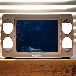 May include: A vintage beige vanity mirror with a dark blue screen and four circular lights. The mirror has a rectangular shape with rounded corners and a small, rectangular base. The lights are evenly spaced around the screen.