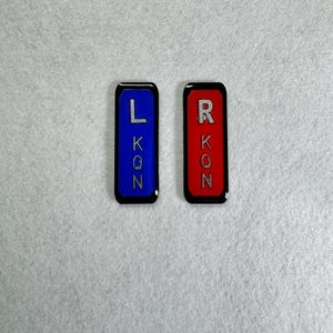 May include: Two rectangular, glossy, black-edged nameplates. One is blue with the letters L, K, Q, and N in silver. The other is red with the letters R, K, O, and N in silver. The nameplates are likely for a product or equipment.