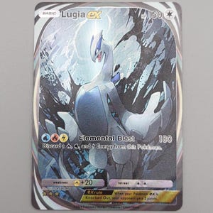 May include: A holographic Pokémon trading card featuring Lugia ex. The card has a silver border and depicts a white and blue Lugia with a dark background. The card text includes "Elemental Blast" and "HP 150".