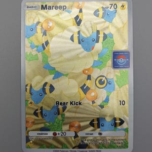 May include: A Mareep Pokémon trading card with a gold foil background. The card features a blue sheep-like creature with yellow accents. Text includes "Mareep", "Rear Kick", and other game-related details. The card is encased in a clear protective sleeve.