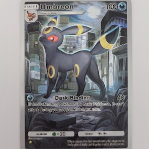 May include: Umbreon Pokémon trading card. The card shows a black creature with yellow details, set against a cityscape. The card features the name "Umbreon", HP 100, and the "Dark Binding" ability. Text on the card describes the creature's abilities.