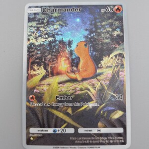 May include: A Pokémon trading card depicting Charmander, an orange lizard-like creature, by a campfire in a forest. The card includes the text "Ember" and "HP 60". The background shows a starry night sky. The card is rectangular.