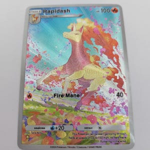 May include: A holographic Rapidash Pokémon card. The card features a running horse-like creature with a pink and yellow body and a fiery mane. The card has text that reads "Fire Mane" and "HP 100". The background is a colorful, abstract design.