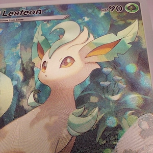 May include: A holographic trading card featuring Leafeon, a green and tan Pok&eacute;mon. The card has a forest background and the HP is 90. The card is from the Eevee evolution line.