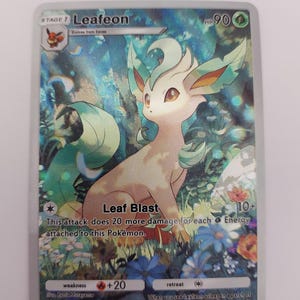 May include: A holographic Pok&eacute;mon trading card featuring Leafeon, a grass-type Pok&eacute;mon. The card has a colourful background with flowers and foliage. The text on the card includes "Leaf Blast" and "HP 90".