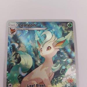 May include: A holographic trading card featuring Leafeon, a Pok&eacute;mon character. The card displays Leafeon with a light brown body, green accents, and the text "Leaf Blast." The card also includes the text "HP 90" and "10+".