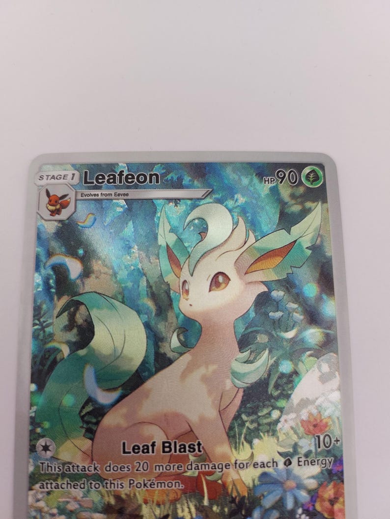 May include: A holographic trading card featuring Leafeon, a Pok&eacute;mon character. The card displays Leafeon with a light brown body, green accents, and the text "Leaf Blast." The card also includes the text "HP 90" and "10+".