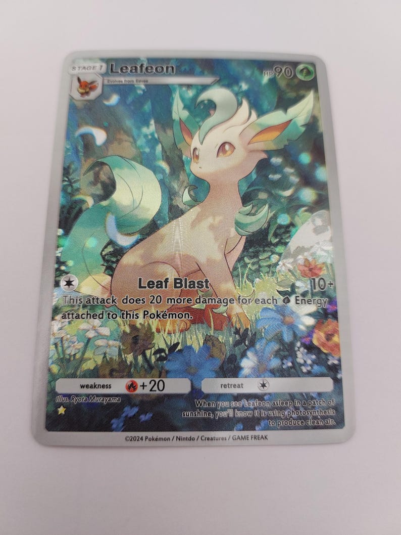 May include: A Pok&eacute;mon trading card featuring Leafeon, a green and tan creature, set against a floral background. The card includes text such as "Leaf Blast" and "+20". The card is from 2024.