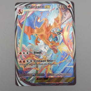 May include: A Charizard EX Pokémon trading card. The card features a vibrant illustration of Charizard in shades of orange and red against a blue and white background. Text includes "Charizard EX", "Slash", and "Crimson Storm".