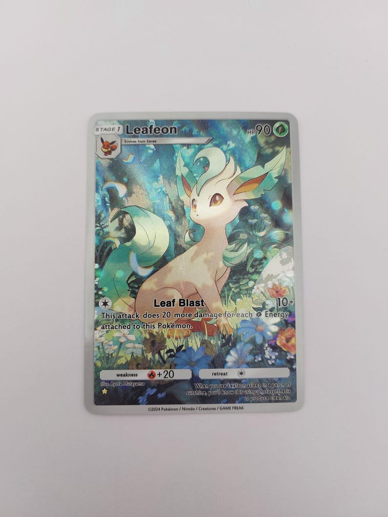 May include: A holographic Leafeon Pok&eacute;mon trading card. The card features a light brown and green creature with a leafy design, set against a background of flowers and foliage. The card has text that reads "Leafeon" and "Leaf Blast".