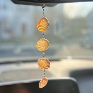 May include: A car charm featuring four natural, fan-shaped seashells in varying sizes, strung together with a silver chain. The shells display a warm, beige color with textured ridges, creating a coastal aesthetic. The charm hangs from a car's rearview mirror.
