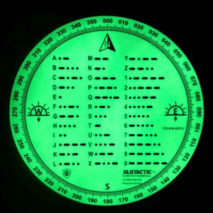 May include: A circular, glow-in-the-dark navigation tool featuring a compass rose with degree markings from 000 to 350. The center displays a Morse code alphabet and numeric chart with black lettering. Text includes GLOTACTIC and ITU-R M.1677-1.