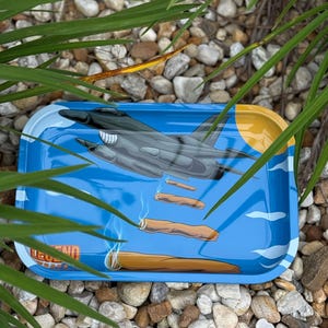 May include: A rectangular blue metal rolling tray with a graphic of a gray jet plane and several lit cigars. The tray has a yellow sun and white cloud design. The word "LEGEND" is printed on the tray.