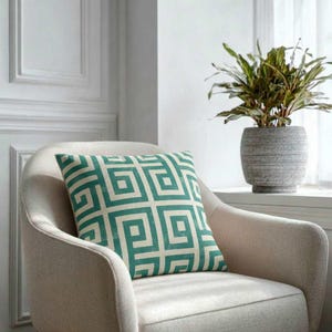 Op de afbeelding: Een vierkant decoratief kussen met een turquoise geometrisch patroon ligt op een beige fauteuil. Het ontwerp van het kussen heeft een herhalend vierkant motief. Een plant in een pot staat op een vensterbank op de achtergrond.