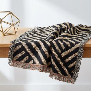 May include: A black and tan throw blanket with a geometric zigzag pattern and fringed edges. The blanket is draped over a wooden surface, showcasing its design and texture. A gold geometric sculpture is in the background.
