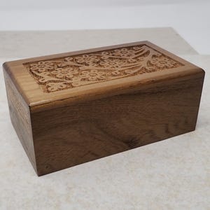 May include: A rectangular wooden box with a floral design carved into the top. The box is made of a light brown wood with a visible grain. The floral pattern is a lighter shade of brown, creating a contrast. The box is closed.