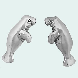 May include: A pair of silver-colored manatee earrings. Each earring is a detailed miniature sculpture of a manatee, with visible flippers, tail, and facial features. The earrings are designed to be worn as a matching set.
