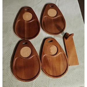 May include: Four teardrop-shaped wooden serving trays, each featuring a circular cork inlay and a small cutout handle at the top. A single rectangular wooden stand with a small angled support piece is positioned to the right of the trays.