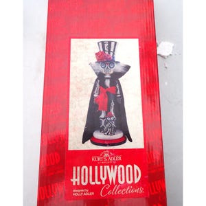 May include: A decorative nutcracker figure featuring a skeleton-themed design with a black cape, a top hat adorned with red roses, and a red bow. The packaging box displays the text Kurt S. Adler Hollywood Collections designed by Holly Adler.