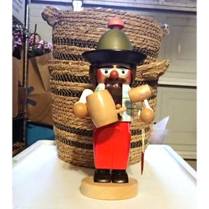 May include: A wooden nutcracker figure wearing a green hat with a red pom-pom and a wide black brim. The figure has a brown beard, red trousers, and a white shirt with a green plaid pattern. It holds a light-colored wooden mug and a small mallet.
