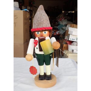 May include: A wooden nutcracker figure wearing a grey felt pointed hat with a red rope trim. The figure has a white shirt with a red heart detail, green shorts, and black shoes. It holds a yellow and green beer mug and has a red tag attached.