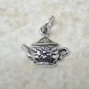 May include: Silver charm in the shape of a teapot with a floral design. The teapot has a lid, a spout, and a handle. The charm is suitable for jewelry making.