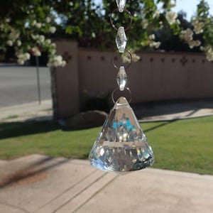 May include: A clear, faceted crystal prism hanging from a chain of small, clear crystals and silver rings. The prism is shaped like a truncated pyramid, reflecting light and creating a rainbow effect. The background is a blurred outdoor scene.