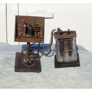 May include: A vintage telegraph apparatus featuring a dark wood base and upright panel with brass components and a black coil. It is connected by coiled metal wires to a glass battery cell container with two black rods mounted on a wooden base.