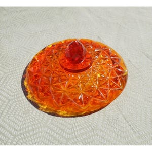 May include: A round, vibrant orange glass lid featuring a geometric starburst pattern across its surface. The lid is topped with a central, faceted, diamond-shaped knob that reflects light, creating a bright and translucent appearance throughout.