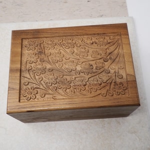 May include: A rectangular wooden box with a floral design carved into the lid. The box is made of light brown wood and has a smooth, polished finish. The floral pattern is detailed and intricate.