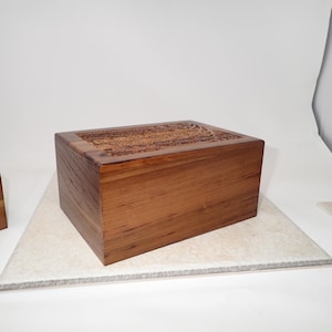 May include: A rectangular wooden box with a carved design on the top. The box is made of a warm brown wood and sits on a light-colored surface. The carved design appears to be floral or ornamental.