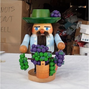 May include: A wooden nutcracker figure wearing a green hat with purple grapes, a light blue jacket, and a dark brown beard. The figure holds clusters of green and purple grapes in each hand and stands behind a wooden bucket on a round base.