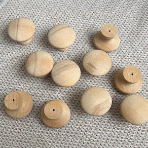 May include: A collection of ten unfinished wooden cabinet knobs. The knobs are round with a natural wood grain finish. Some knobs have a small, cylindrical base with a pre-drilled hole for mounting. The knobs are arranged on a patterned fabric.