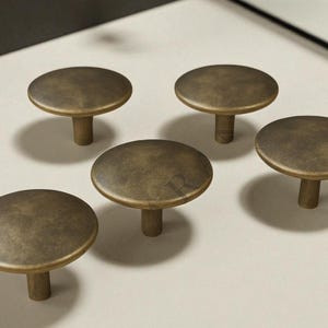 May include: Five antique brass cabinet knobs with a flat, circular top and a short, cylindrical stem. The knobs are arranged on a light-colored surface, casting soft shadows. The hardware has a vintage, aged appearance.