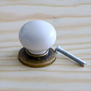 Könnte beinhalten: Ein weißer Keramik-Schrankknopf mit runder Form, auf einem bronzefarbenen Sockel. Eine silberne Schraube befindet sich neben dem Knopf. Die Hardware ist für Möbel und Heimdekoration konzipiert.