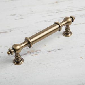 Puede incluir: Un tirador de armario de color bronce, con una sección central cilíndrica y extremos decorativos. La ferretería se muestra sobre una superficie de madera blanca envejecida, resaltando su estética vintage. El tirador mide unos 15 cm de largo.