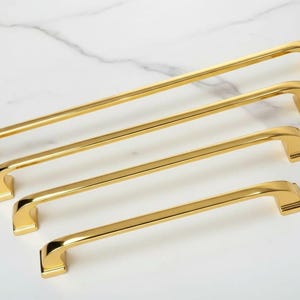 Könnte beinhalten: Vier goldfarbene Schrankgriffe in verschiedenen Längen. Jeder Griff hat eine rechteckige Basis und einen gebogenen Griff. Die Griffe sind diagonal angeordnet und zeigen ihre polierte Oberfläche und ihr Design. Ideal für Küchen- oder Badezimmerschränke.