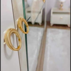 May include: Close-up of a white cabinet door with two gold ring-shaped handles. The handles are mounted on the door, reflecting the room's interior, including a mirror and a bedside table.