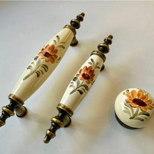 Könnte beinhalten: Zwei cremefarbene Schrankgriffe und ein passender Knauf. Jeder Griff hat ein Blumendesign mit orangefarbenen und braunen Blumen, grünen Blättern und goldfarbener Hardware. Der Knauf hat ein ähnliches Blumendesign.