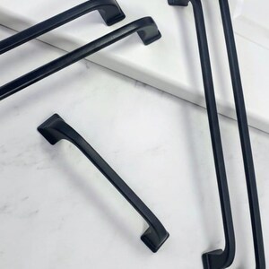 May include: Several black cabinet pulls in various sizes are arranged on a white surface. The pulls have a sleek, modern design with a curved handle and square mounting plates. The hardware is made of a matte black metal.