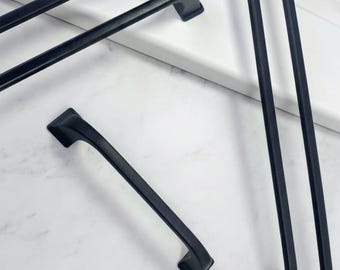 Może przedstawiać: Kilka czarnych uchwytów do szafek w różnych rozmiarach ułożonych na białej powierzchni. Uchwyty mają elegancki, nowoczesny design z zakrzywionym uchwytem i kwadratowymi płytkami montażowymi. Okucia wykonane są z matowego czarnego metalu.