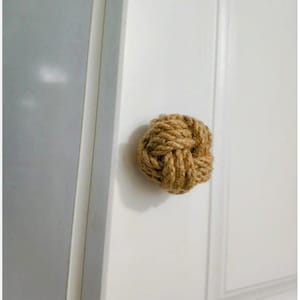 Pode incluir: Um puxador de porta decorativo feito de corda natural trançada, assemelhando-se a um nó náutico. O puxador tem uma cor castanha quente e está montado numa porta branca. O design adiciona um elemento rústico e texturizado à porta.