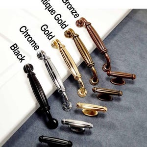 以下が含まれることがあります： 黒、クロム、ゴールド、アンティークゴールド、ブロンズなど、さまざまな仕上げのキャビネットハードウェアのセレクション。画像には、さまざまなデザインと色を示す、いくつかのスタイルのハンドルとノブが表示されています。