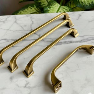 May include: A collection of antique brass cabinet pulls in various sizes. The pulls have a curved design with square bases and are arranged on a marble surface. The brass finish gives them a vintage look.
